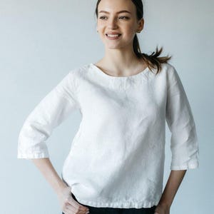 May include: A white long-sleeved top with a round neckline and a cinched hemline. The garment features three-quarter length sleeves and a relaxed fit, constructed from a textured fabric that sits at the waistline over dark-coloured trousers.