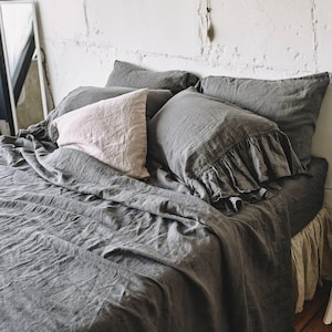 May include: A charcoal gray bedding set featuring a duvet cover and two matching pillowcases with ruffled edges. A single light beige pillowcase is placed on top of the dark gray bedding. The fabric has a visible textured weave throughout the set.