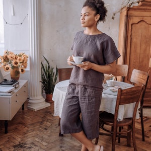 May include: A woman wearing a grey linen jumpsuit and pink sliders stands in a room with a wooden floor and a white table. She is holding a white cup and saucer in her hands.