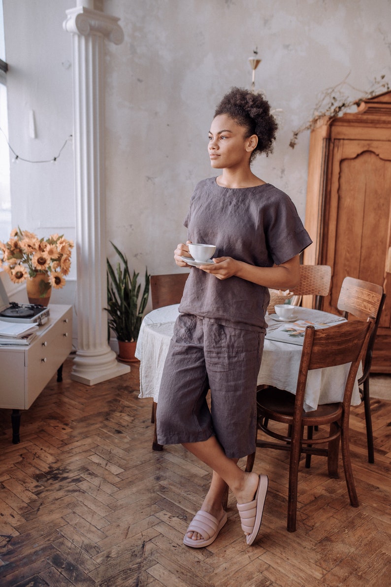 May include: A woman wearing a grey linen jumpsuit and pink sliders stands in a room with a wooden floor and a white table. She is holding a white cup and saucer in her hands.