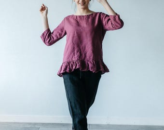 May include: A dark magenta long-sleeved top with a round neckline and a ruffled hemline, paired with black ankle-length trousers. The top features a loose fit and three-quarter length sleeves, while the trousers have a simple, straight-leg cut.