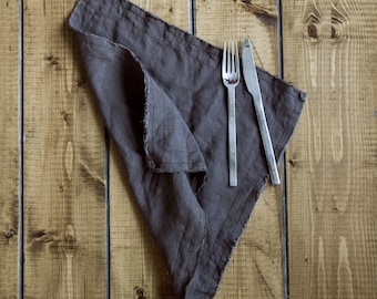 May include: A dark charcoal gray fabric napkin with a frayed edge, folded into a triangular shape. A stainless steel fork and a stainless steel knife are placed side by side on top of the fabric napkin, aligned vertically with the folded edge.