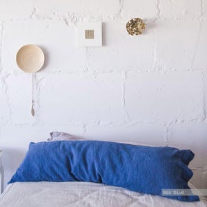May include: A long, rectangular sea blue pillowcase made of textured fabric, positioned horizontally on a bed. The pillowcase features a deep, solid blue color and a visible weave, resting against a white, block-patterned wall with decor.