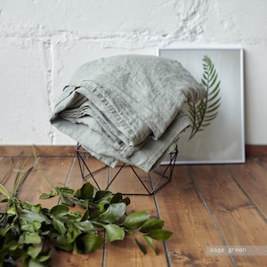 May include: A sage green fabric textile with a visible woven texture, folded and draped over a black wire geometric frame. The fabric displays a soft, muted green tone and natural creases throughout its surface, showcasing a simple, functional design.