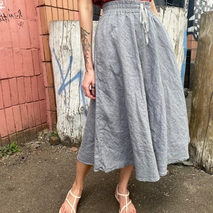 K&ouml;nnte beinhalten: Ein grauer Leinenrock mit einem Kordelzugbund und einer lockeren Passform. Der Rock ist knielang und hat einen leicht ausgestellten Saum. Das Model tr&auml;gt den Rock zu einem rostfarbenen Tanktop und wei&szlig;en Sandalen.