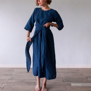 May include: A sea blue midi-length dress with three-quarter length sleeves, a round neckline, and a wrap-style waist featuring a long, integrated fabric tie belt. The garment is made of a solid-colored textile and falls to a loose, flowing skirt.