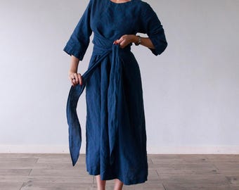 May include: A sea blue midi-length dress with three-quarter length sleeves, a round neckline, and a wrap-style waist featuring a long, integrated fabric tie belt. The garment is made of a solid-colored textile and falls to a loose, flowing skirt.