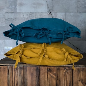 May include: Two linen pillow covers, one teal and one mustard yellow, stacked on a dark wooden surface. Each pillow has tie closures on the side. The pillows are rectangular and appear to be filled.