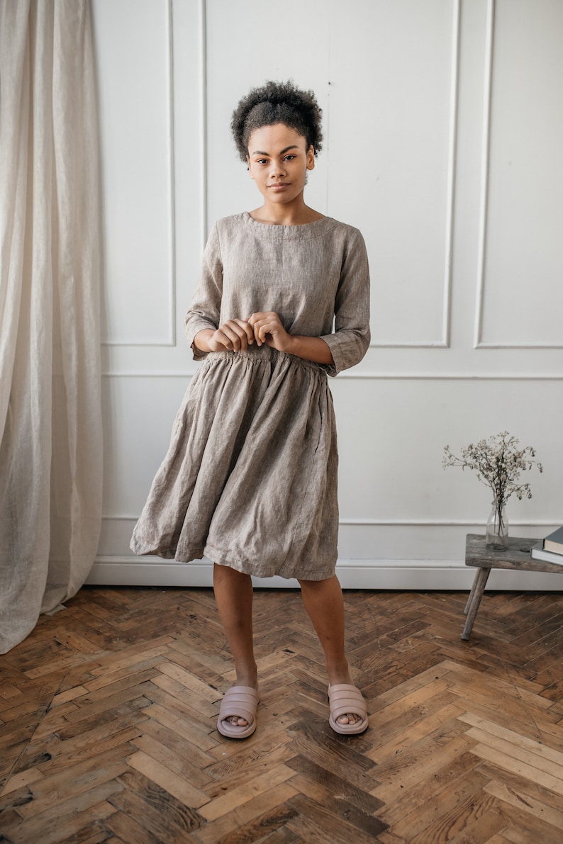 May include: A woman wearing a light brown linen dress with a gathered skirt. She is standing on a wooden floor with a white wall in the background.