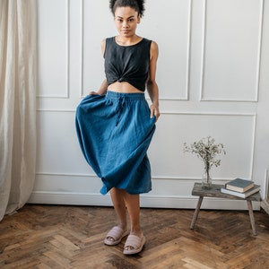 Pu&ograve; includere: Una donna indossa un top nero senza maniche con un nodo frontale e una gonna blu con coulisse. Indossa anche delle pantofole rosa. La stanza ha una parete bianca e un pavimento in legno.