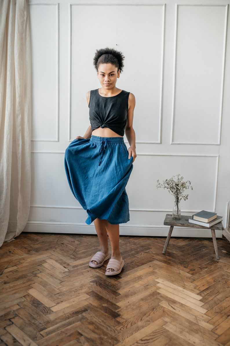 Pu&ograve; includere: Una donna indossa un top nero senza maniche con un nodo frontale e una gonna blu con coulisse. Indossa anche delle pantofole rosa. La stanza ha una parete bianca e un pavimento in legno.
