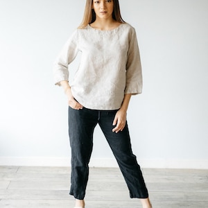 May include: A beige long-sleeved top with a round neckline and three-quarter length sleeves paired with black cropped trousers and light-colored open-toe sandals. The outfit consists of a neutral-toned blouse and dark pants with a simple design.