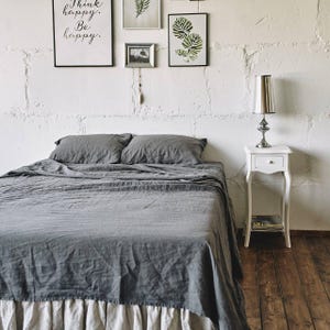 May include: A bed with dark gray linen bedding, including two pillows and a flat sheet, featuring a light beige ruffled bed skirt. Beside the bed stands a white wooden nightstand with a silver metallic lamp topped with a conical shade.