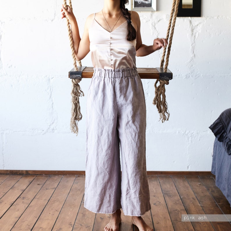 May include: A woman wearing a light pink satin camisole top and wide-leg, high-waisted linen pants in a pink ash color. The pants have an elastic waistband. The model is standing near a wooden swing with rope supports.