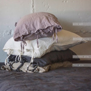 May include: A stack of four linen pillowcases in various shades of gray, including pink ash, antique white, charcoal gray, and flax gray. The pillowcases have ties at the corners.