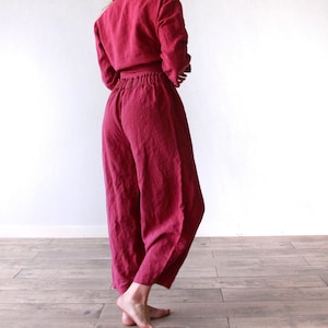 May include: A deep magenta jumpsuit featuring long sleeves and a cinched waistline. The garment has a gathered elastic waistband and wide-leg trousers that fall to the lower calf, crafted from a textured fabric with visible weave details.