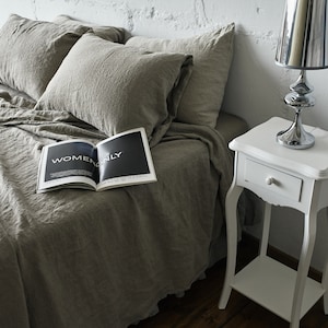 May include: A white bedside table with a drawer and a lower shelf. A silver lamp with a white lampshade sits on the table. A magazine with the text "Women Only" is on a bed with linen bedding.