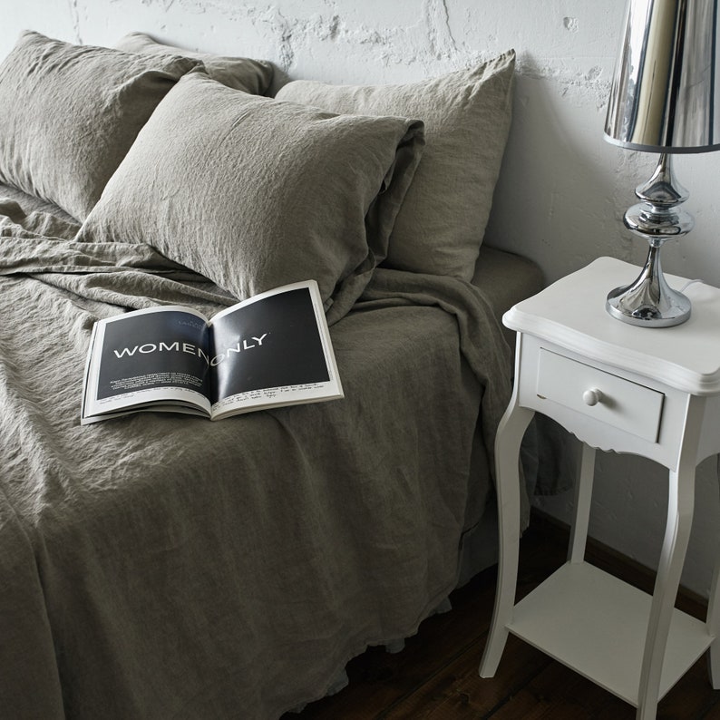 May include: A white bedside table with a drawer and a lower shelf. A silver lamp with a white lampshade sits on the table. A magazine with the text "Women Only" is on a bed with linen bedding.