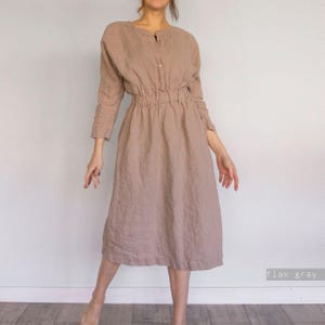 May include: A long-sleeved midi dress in a solid flax grey colour. The dress features a round neckline with a partial button-down placket, a gathered elasticised waistline, and a loose-fitting skirt that falls below the knees in a simple silhouette.