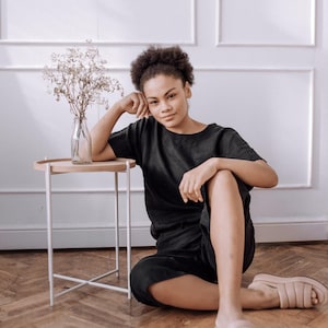 May include: A black short-sleeved top and matching shorts set paired with beige padded slide sandals. The outfit features a relaxed fit with a simple round neckline. The ensemble is worn with a neutral aesthetic, focusing on the solid black fabric.
