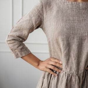 May include: A brown linen dress with a scoop neck and 3/4 sleeves. The dress has a gathered waist and a flowing skirt.