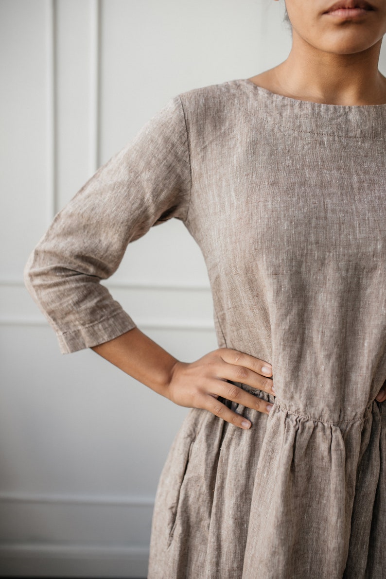 May include: A brown linen dress with a scoop neck and 3/4 sleeves. The dress has a gathered waist and a flowing skirt.