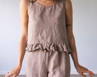 Puede incluir: Una blusa sin mangas en color gris lino con cuello redondo y un detalle de volante en el dobladillo de la cintura. La prenda tiene una apariencia texturizada y se combina con pantalones a juego en el mismo tono gris lino y tejido.