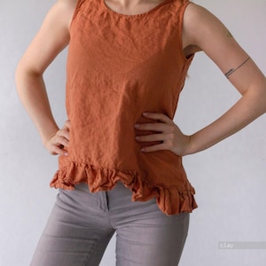 May include: A sleeveless top in a solid burnt orange color featuring a round neckline and a ruffled hemline. The garment is constructed with a textured fabric and falls to the hip, providing a simple silhouette with a gathered bottom edge.