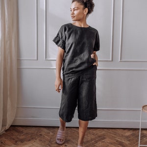 Peut inclure: Une personne portant un haut en lin noir et un short en lin assorti. La personne porte des tongs roses et se tient debout sur un sol en bois.
