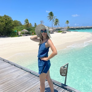 以下が含まれることがあります： 青いドレスを着て、つば広の麦わら帽子をかぶった女性が、ターコイズブルーのラグーンを見下ろす木製の桟橋に立っています。ビーチは白い砂浜で、背景にはヤシの木があります。