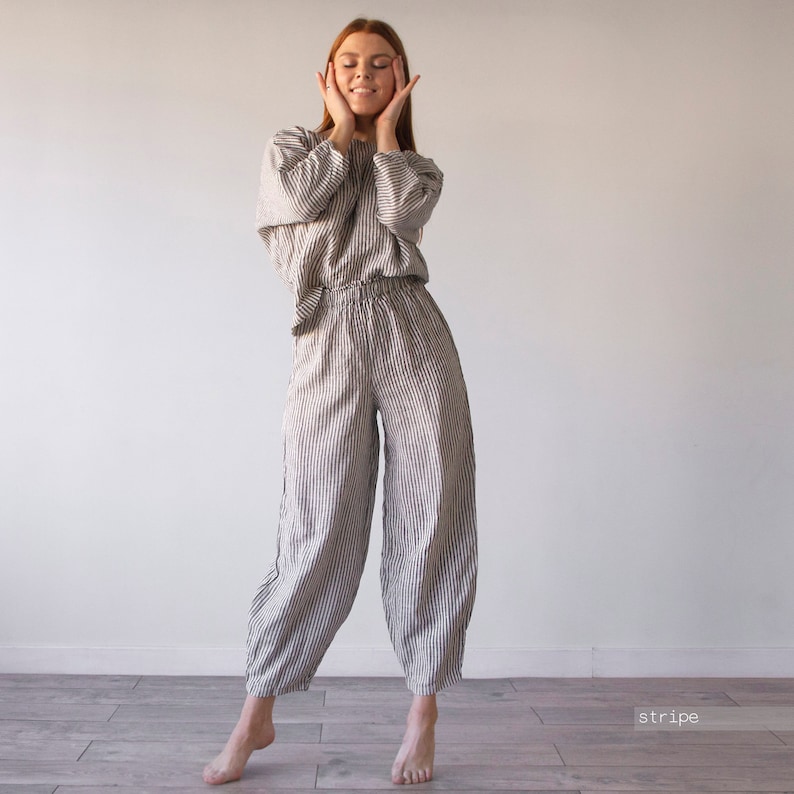 Pode incluir: Uma mulher vestindo um macac&atilde;o de linho listrado. O macac&atilde;o &eacute; de cor cinza claro com listras pretas. A mulher est&aacute; de p&eacute; com as m&atilde;os nas bochechas e os olhos fechados.