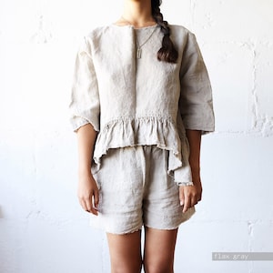 May include: A flax gray two-piece set consisting of a top and shorts. The top features a round neckline, three-quarter length sleeves, and a ruffled hem with a raw edge. The matching shorts have a simple cut and a raw hem. The fabric has a textured appearance.