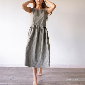 May include: A sleeveless sage green midi dress featuring a round neckline and a gathered waistline that flows into a relaxed, loose-fitting skirt. The garment is made of a textured fabric and reaches below the knees, with the text sage green visible.