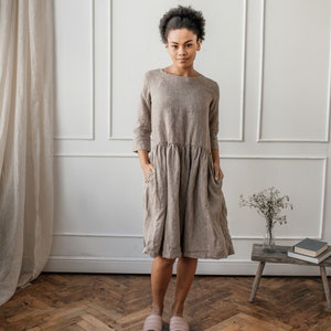 May include: A woman wearing a brown linen dress with 3/4 sleeves and pockets. She is standing on a wood floor with a white wall and a small wooden table behind her.