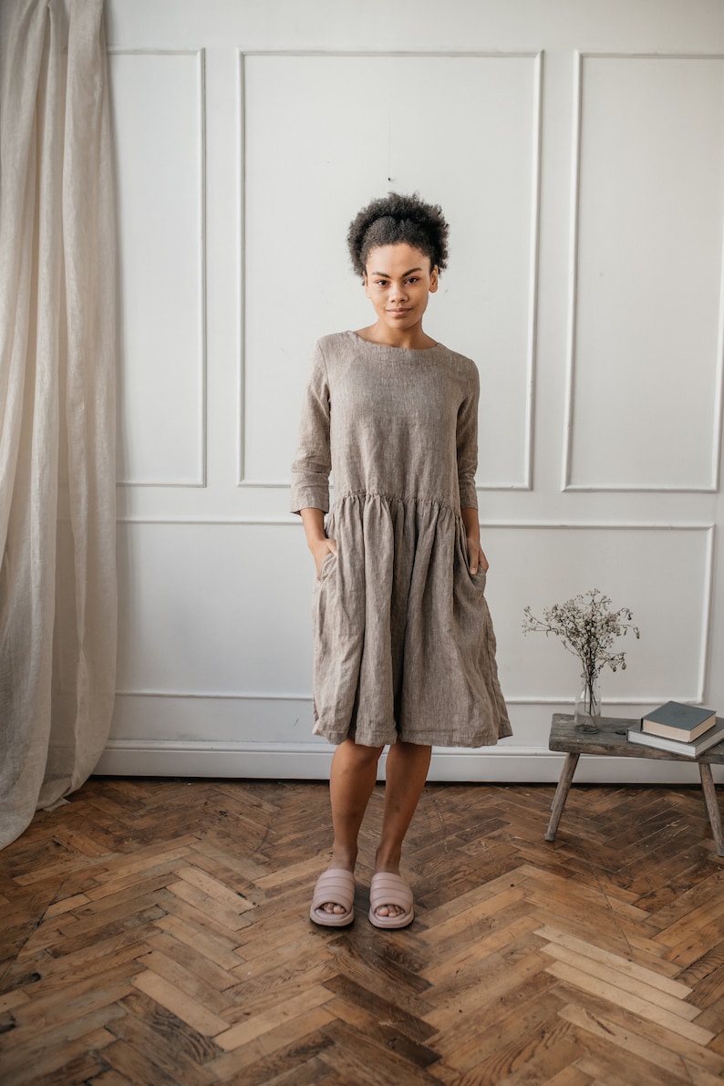 May include: A woman wearing a brown linen dress with 3/4 sleeves and pockets. She is standing on a wood floor with a white wall and a small wooden table behind her.