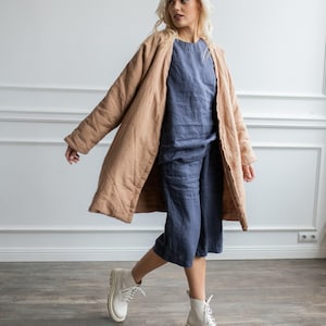 May include: A long, quilted tan coat worn over a blue linen top and matching blue culottes, paired with white lace-up boots. The coat features a relaxed fit with long sleeves, while the blue set consists of a sleeveless top and cropped pants.