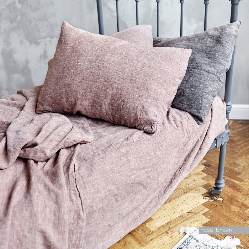 May include: A rose brown linen bed sheet and pillowcases. The bed is made with a light pink linen sheet and two pillows, one with a gray linen pillowcase and one with a rose brown linen pillowcase.