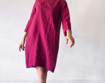 Puede incluir: Un vestido magenta hasta la rodilla con cuello redondo y mangas tres cuartos. La prenda presenta un acabado de tela texturizada y una silueta holgada de corte recto. La palabra marsala está impresa en la esquina inferior derecha.