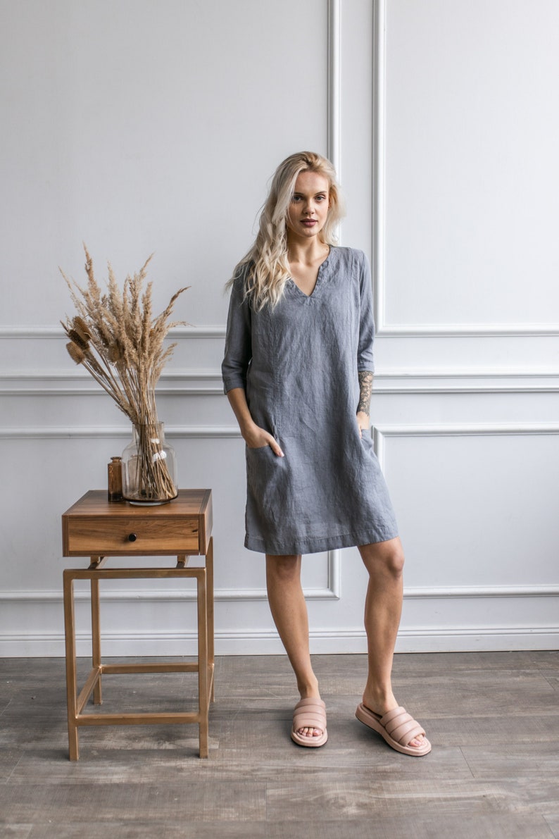 May include: A woman wearing a gray linen dress with a v-neck and pockets. She is standing in front of a white wall with a wooden side table and a vase of dried flowers.