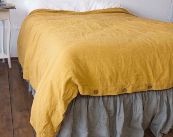 May include: A mustard yellow duvet cover with a button closure along the bottom edge, layered over a light gray ruffled bed skirt. The bedding is draped over a mattress, showcasing a textured fabric finish and a two-tone color combination.
