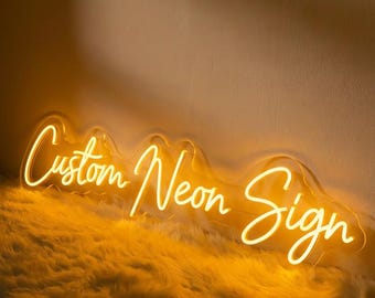 May include: A glowing neon sign in a warm yellow hue, displaying the words "Custom Neon Sign" in a cursive font. The sign is mounted on a fluffy, textured surface, creating a soft contrast with the bright light.
