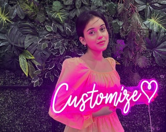 May include: A bright pink neon sign featuring the word Customize in a cursive script font followed by a heart symbol. The sign is illuminated and emits a vibrant glow, with a visible white power cord extending from the right side of the frame.
