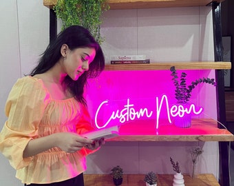May include: A pink neon sign with the text "Custom Neon" in white. The sign is on a wooden shelf with a potted plant and a book.