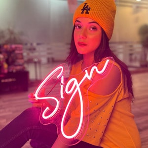May include: A bright pink neon sign in cursive script that reads Sign, held by a person wearing a mustard yellow knit beanie with a dark blue LA logo and a matching mustard yellow cold-shoulder top with silver studs along the sleeves.