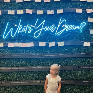 May include: A young child stands in front of a wall with the neon sign "What's Your Dream?". The child wears a light-colored top with a ruffled collar and striped culottes. The wall is covered in green foliage and small notes.