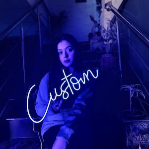 May include: A bright neon sign displaying the word Custom in a cursive script font. The sign emits a vibrant blue light, illuminating the surrounding area with a cool-toned glow. The lettering is connected in a single continuous line.