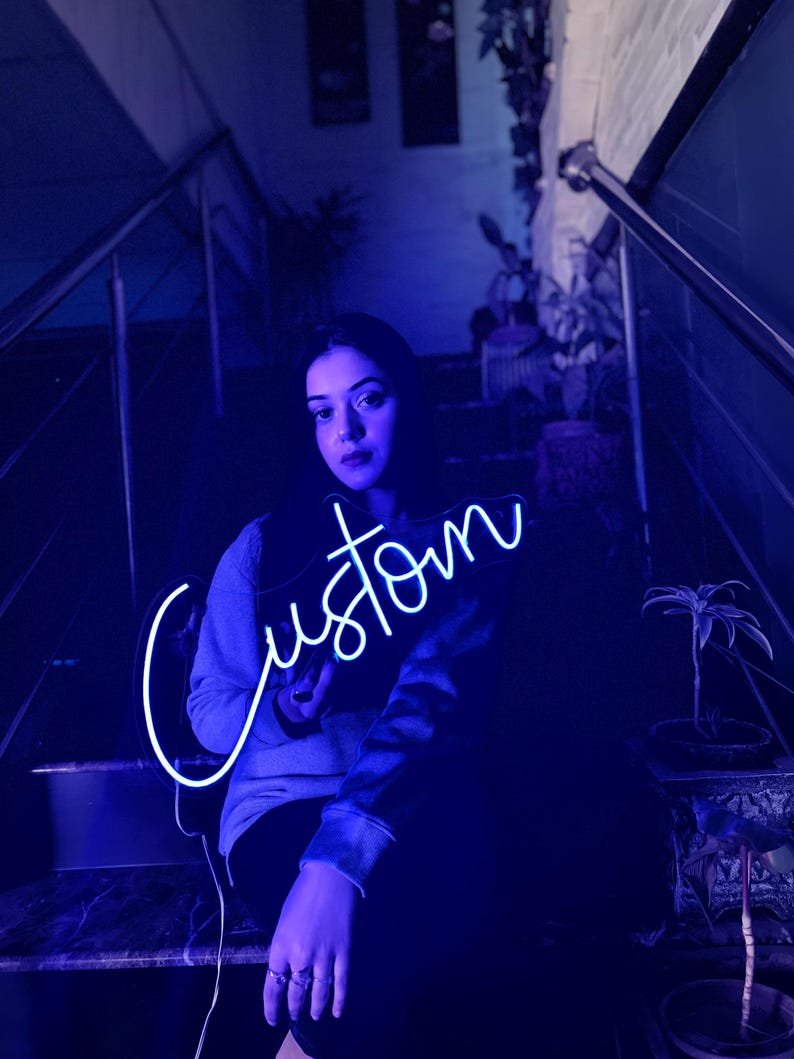 May include: A bright neon sign displaying the word Custom in a cursive script font. The sign emits a vibrant blue light, illuminating the surrounding area with a cool-toned glow. The lettering is connected in a single continuous line.