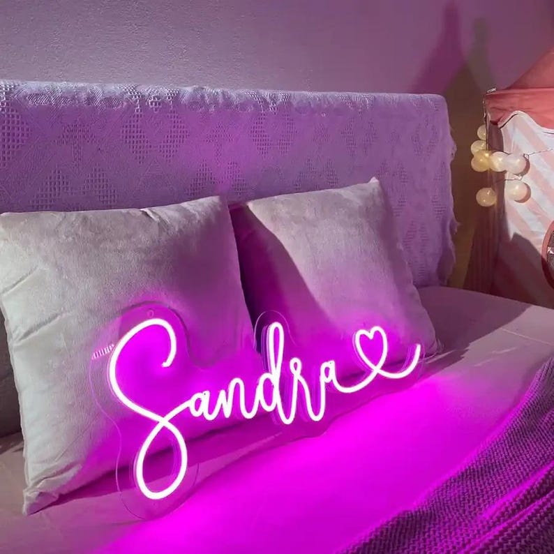 May include: A neon sign in the shape of the name "Sandra" in a cursive font, with a heart at the end. The sign glows bright pink and is mounted on a clear backing. The sign is placed on a bed with pillows.