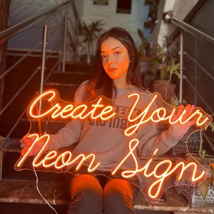 May include: A custom neon sign with orange glowing tubing shaped into the cursive text Create Your Neon Sign. The sign is mounted on a clear, irregularly shaped acrylic backing and includes a white power cord extending from the bottom left corner.