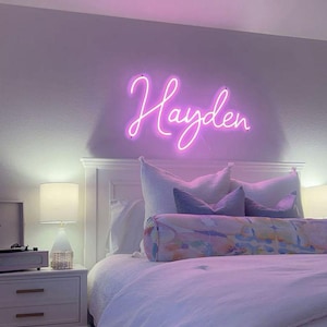 May include: A bedroom scene featuring a white bed with a white comforter and pillows. Above the bed, a neon sign spells out the name "Hayden" in a cursive font. A bedside table with a lamp and a record player is visible.